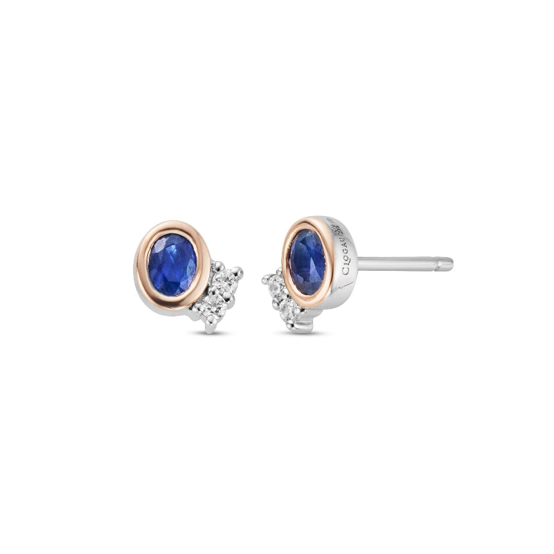 A pair of stud earrings featuring oval blue sapphires in rose gold bezels, accented with small clear stones on a white background.