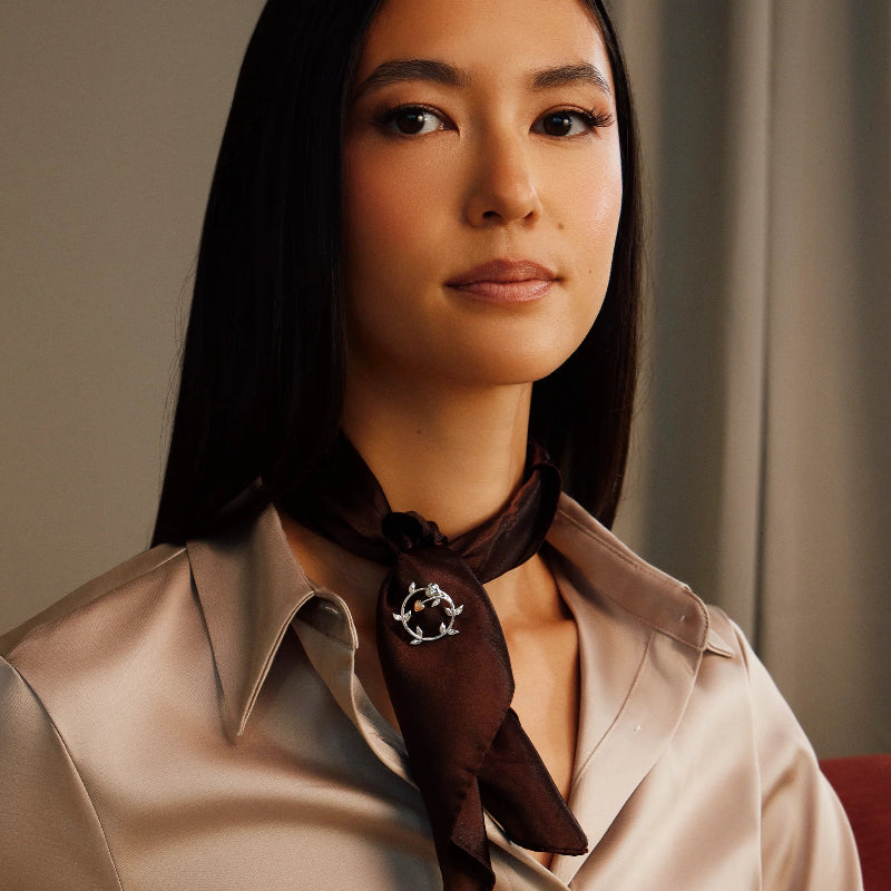 A woman with long dark hair wearing a beige satin shirt and a dark brown silk neck scarf with a silver circular logo, photographed in a warm, softly lit indoor setting.