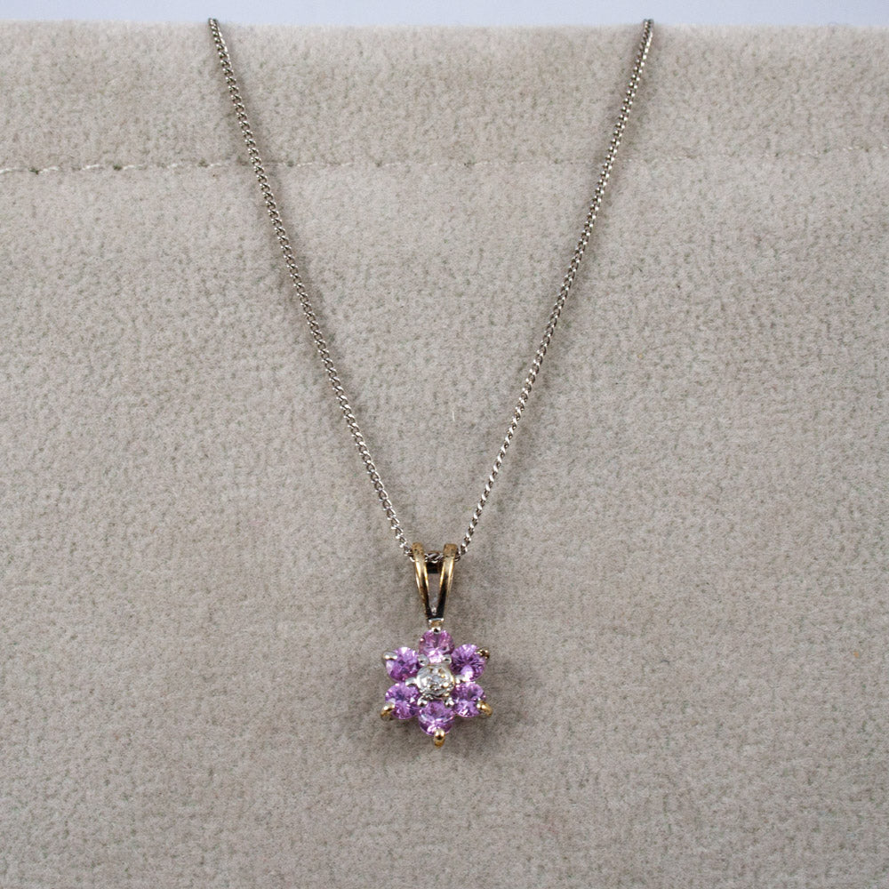 A silver chain necklace with a flower-shaped pendant featuring pink sapphire stones and a central diamond gemstone, displayed on a textured beige jewelry stand.