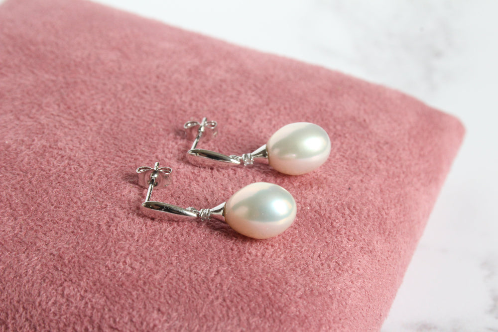 A pair of 9ct white gold drop earrings with diamond and with large white pearl laying on pink cushion - carathea & jools