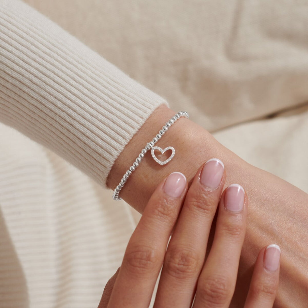 A delicate silver chain bracelet with a small open heart charm, worn on a wrist with a cream ribbed knit sleeve and manicured nails visible.