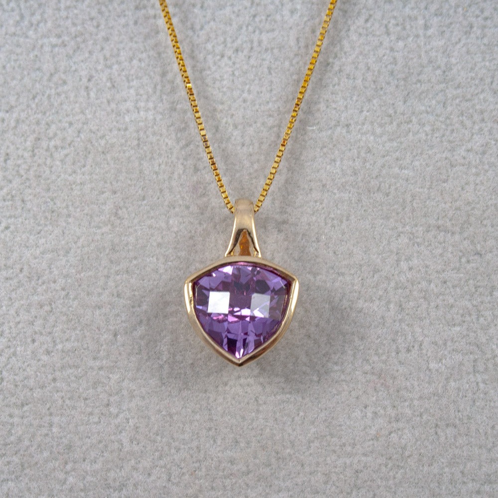A gold pendant necklace featuring a faceted shield-shaped purple created Alexandrite gemstone, displayed on a textured light gray background.