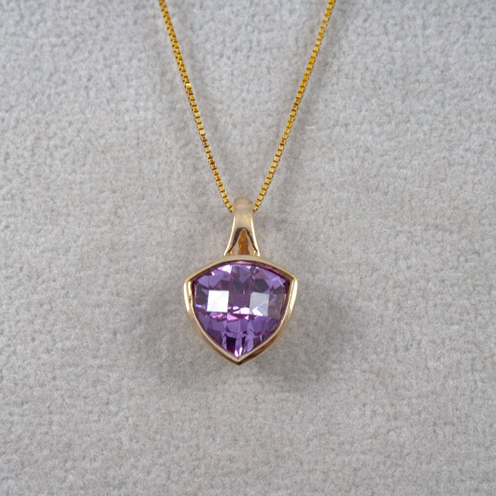 A gold pendant necklace featuring a faceted shield-shaped purple created Alexandrite gemstone, displayed on a textured light gray background.