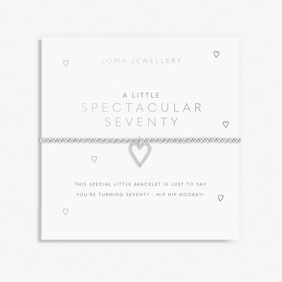 A silver bracelet with a heart-shaped charm, presented on a white card from Joma Jewellery that reads 'A Little Spectacular Seventy'.