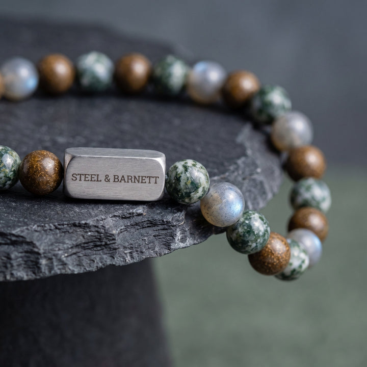 A beaded stretch bracelet with polished stone beads in green, brown, and white, featuring a rectangular silver-tone metal charm engraved with 'STEEL & BARNETT'.