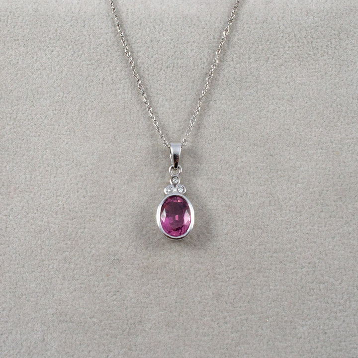A white gold chain necklace with an oval pink tourmaline gemstone with three diamonds pendant, displayed on a textured light gray background.