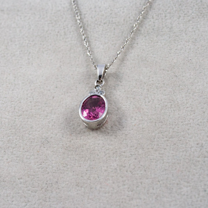 A white gold chain necklace with an oval pink tourmaline gemstone with three diamonds pendant, displayed on a textured light gray background.
