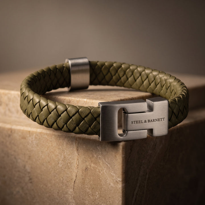 An olive green braided leather bracelet with a brushed silver-tone metal H-shaped clasp engraved with 'STEEL & BARNETT', resting on a stone pedestal.