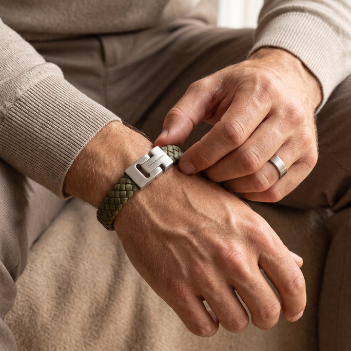 A close-up of a man's hands adjusting an olive green braided leather bracelet with a silver metal clasp on his wrist, wearing a beige sweater and trousers.