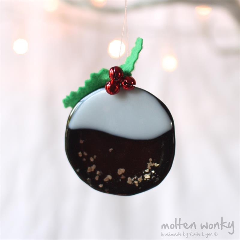 A handmade mottled glass pudding ornament hanging from a thin string, featuring a glossy dark brown body with a white top section, red berry accents, and green leaf details, set against a soft white fabric background with warm bokeh lights.