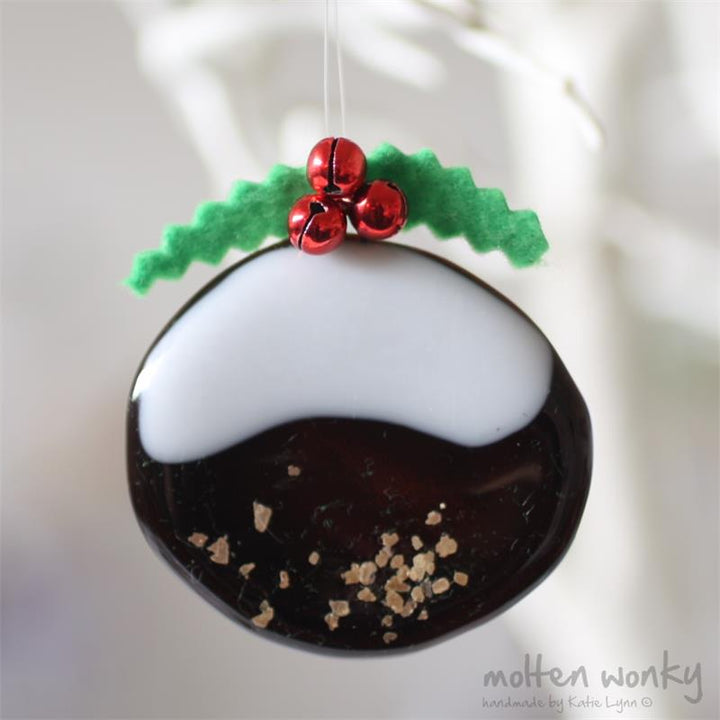 A handmade felt Christmas pudding ornament hanging from a thin string. The ornament features a dark brown lower half with gold glitter flakes, a white upper half, green felt holly leaves, and three shiny red felt berries.