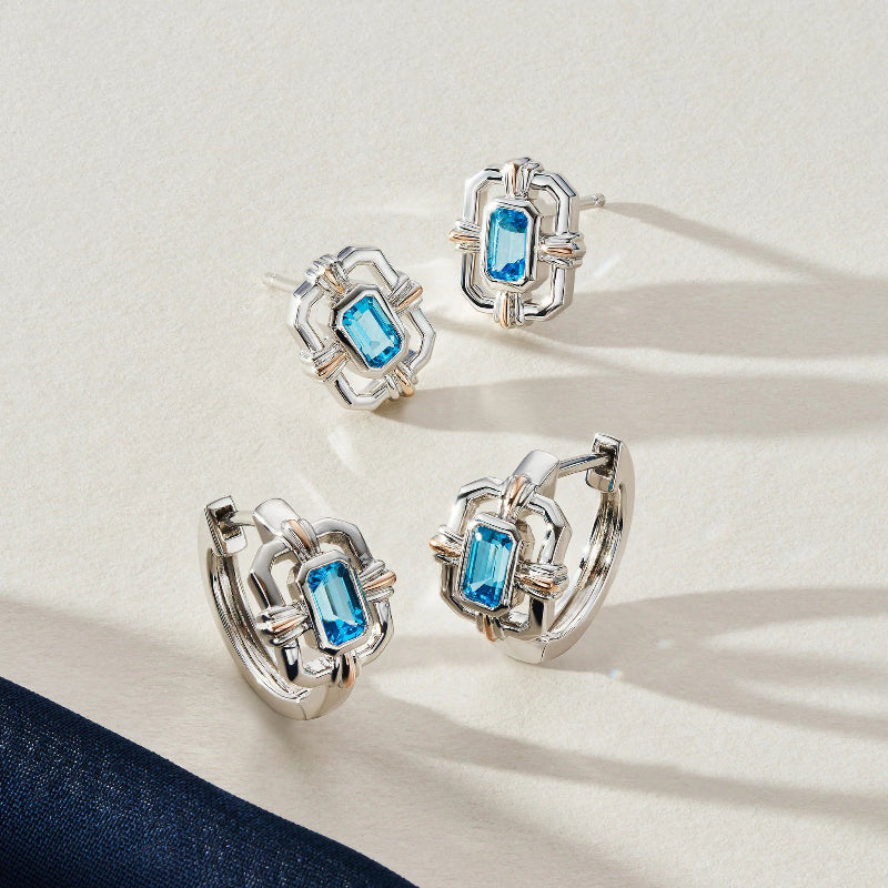 Four silver-toned hoop earrings with rectangular blue gemstones, arranged on a light cream surface with soft shadows and a dark navy fabric corner at the bottom left.