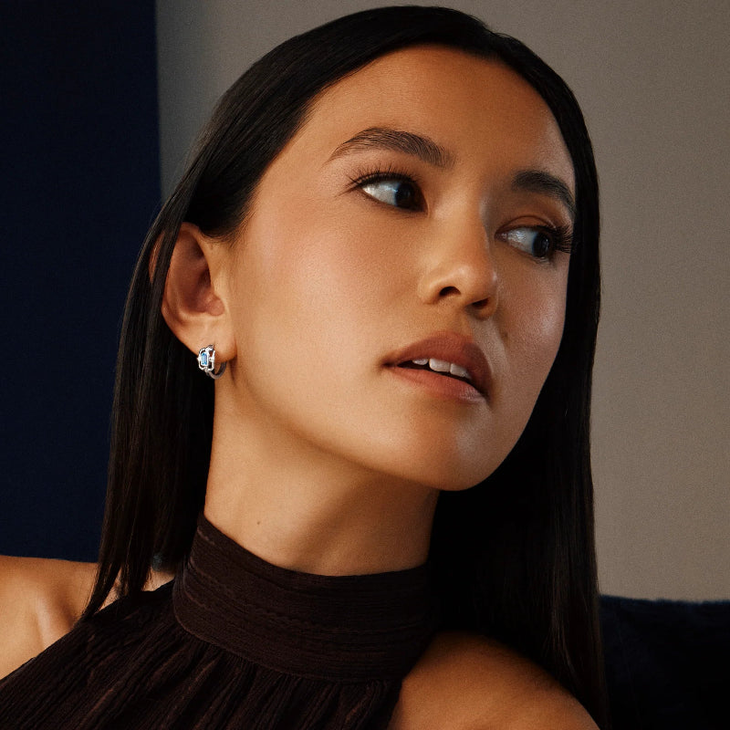 A close-up portrait of a woman with long dark hair wearing a small silver stud earring, set against a dark navy and gray background.