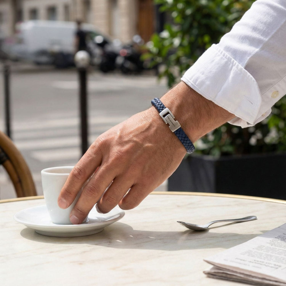 Man with blue leather bracelet holding a coffee cup on an outdoor table with a city street background