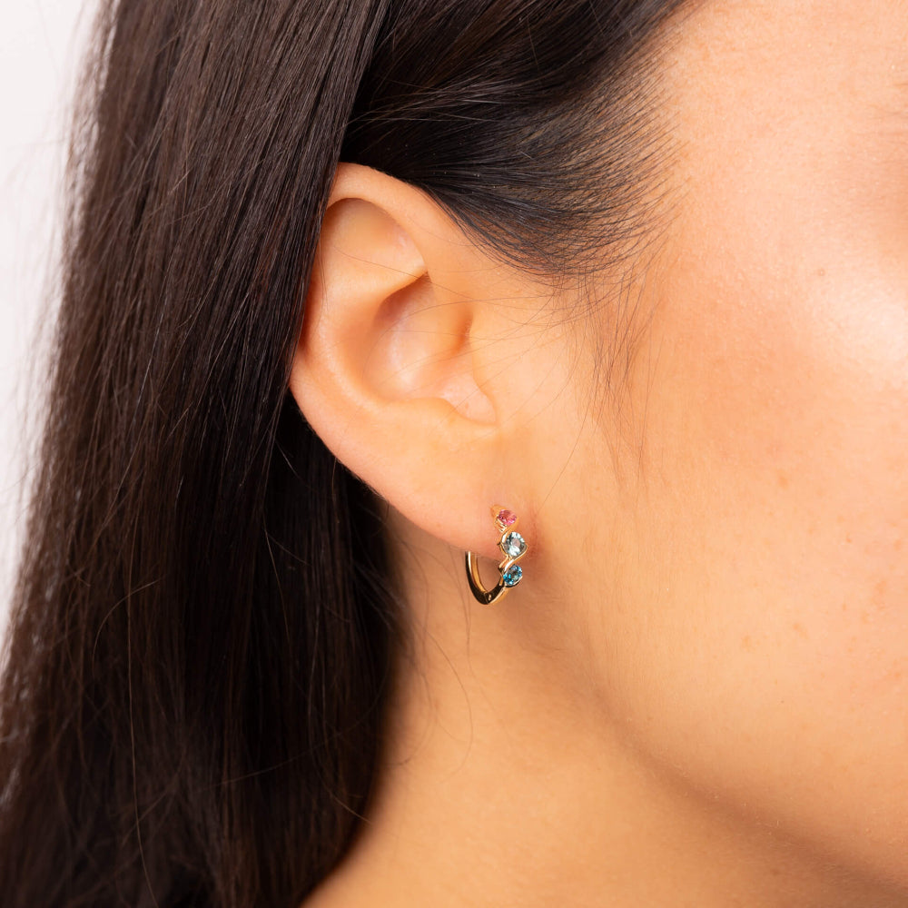 A woman wearing a 9ct yellow gold hoop earring with pink, green and blue stones set in a twisting gold band.