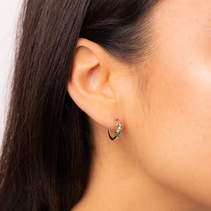 A woman wearing a 9ct yellow gold hoop earring with pink, green and blue stones set in a twisting gold band.
