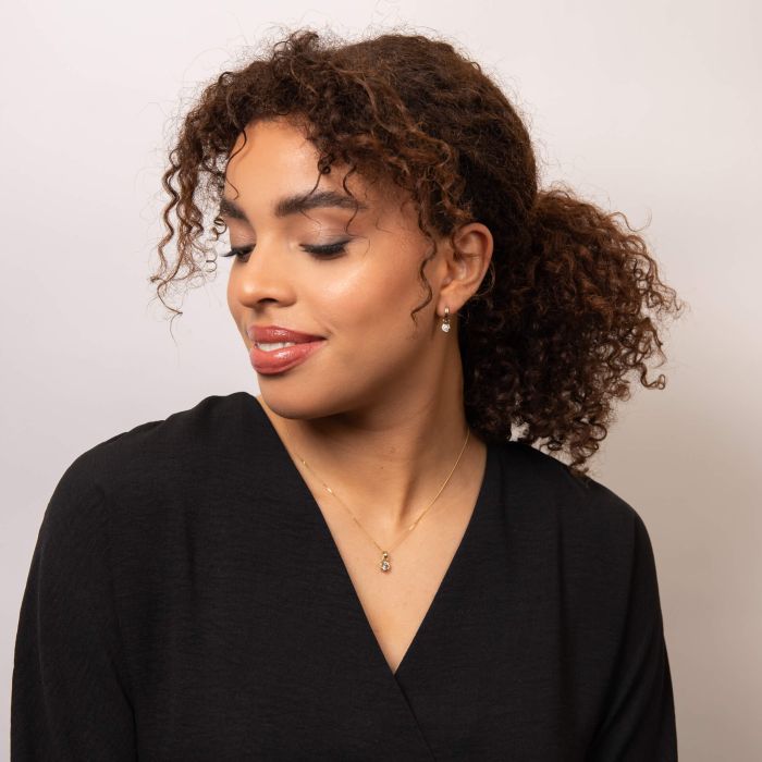 Woman with curly hair styled in a ponytail wearing a black top against a plain background wearing gold hoops and necklace with a diamond hexagonal charm