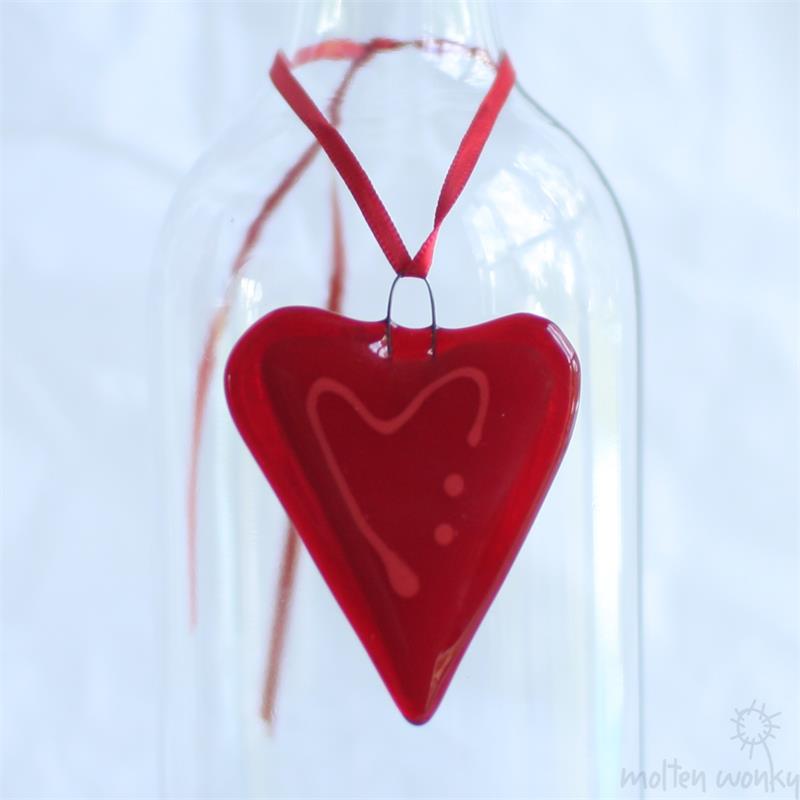 A glossy red heart-shaped glass ornament with a white heart design and three dots, hanging from a red ribbon inside a clear glass bottle against a white background.