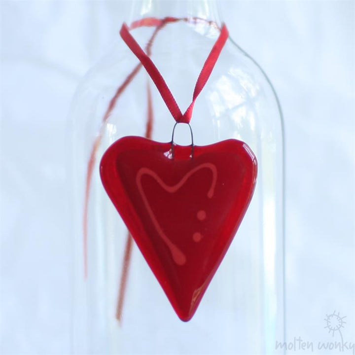 A glossy red heart-shaped glass ornament with a white heart design and three dots, hanging from a red ribbon inside a clear glass bottle against a white background.