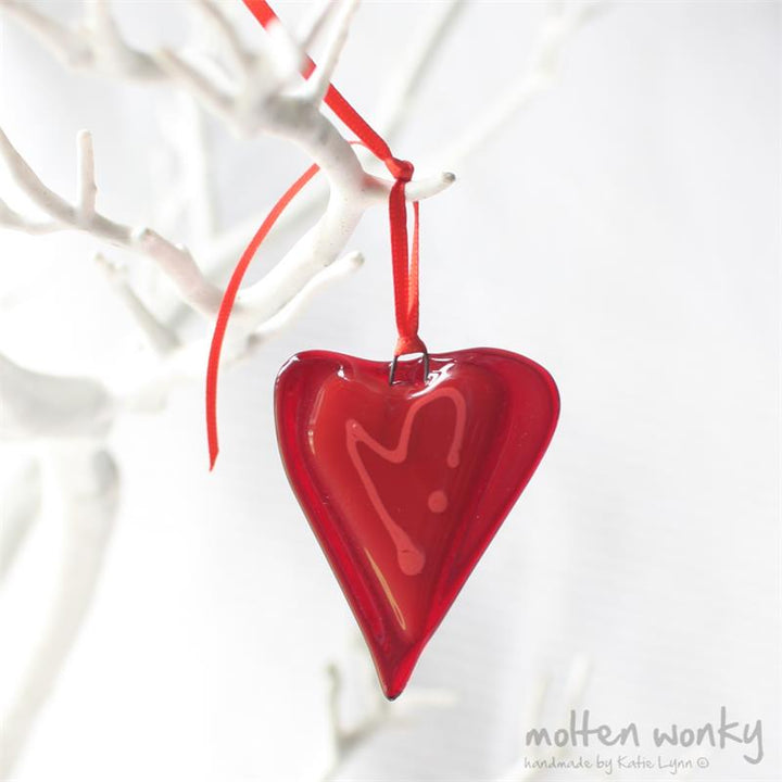 A glossy red heart-shaped glass ornament hanging from a red ribbon, displayed against a white decorative branch backdrop.