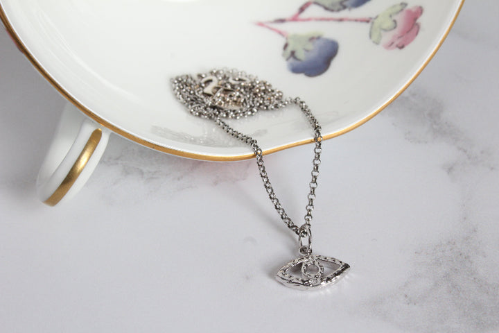 A silver evil eye necklace with a textured pendant draped over a white porcelain plate with gold trim and floral detailing, set on a light marble surface.