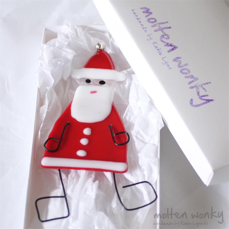 A handmade red and white Santa Claus ornament displayed in its white gift box with tissue paper, next to the brand's white packaging box.
