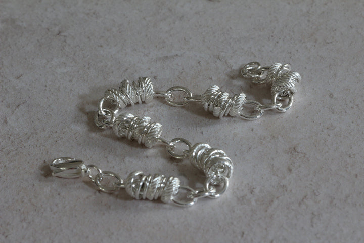 A silver-toned chain accessory with textured spiral links and small oval connectors, laid on a light gray stone-like surface.