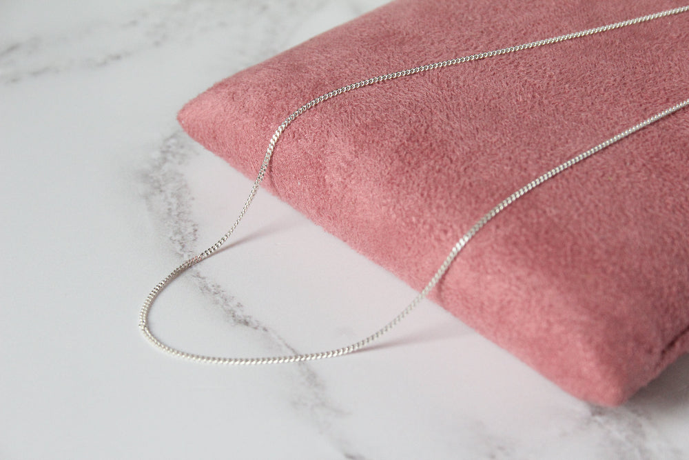 A close-up of a silver chain necklace draped over a dusty pink velvet pouch on a white marble surface.
