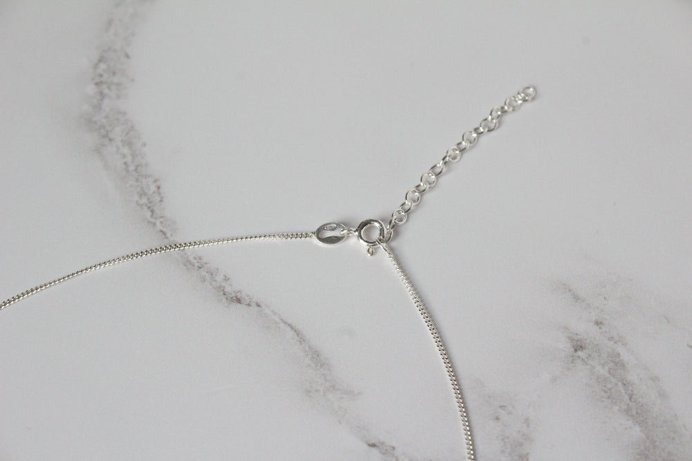 A close-up of a silver curb chain necklace with a bolt clasp and extender chain, laid on a white marble surface - Carathea & Jools