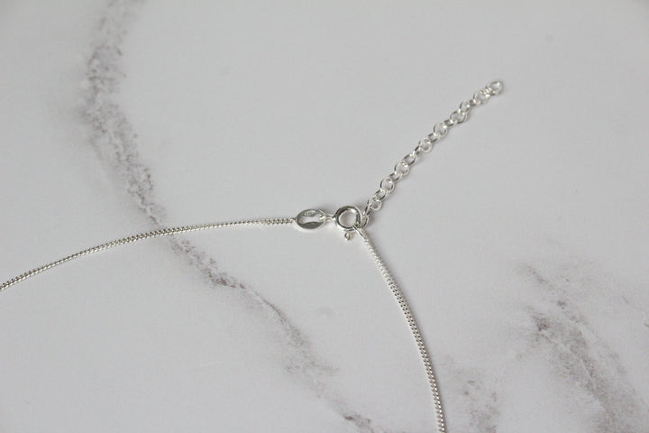 A close-up of a silver curb chain necklace with a bolt clasp and extender chain, laid on a white marble surface - Carathea & Jools