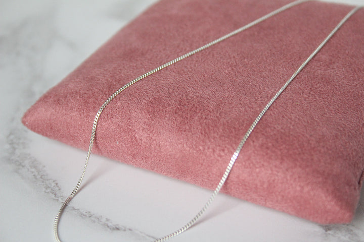 A close-up of a silver curb chain draped over a dusty pink velvet jewelry display cushion on a white marble surface.