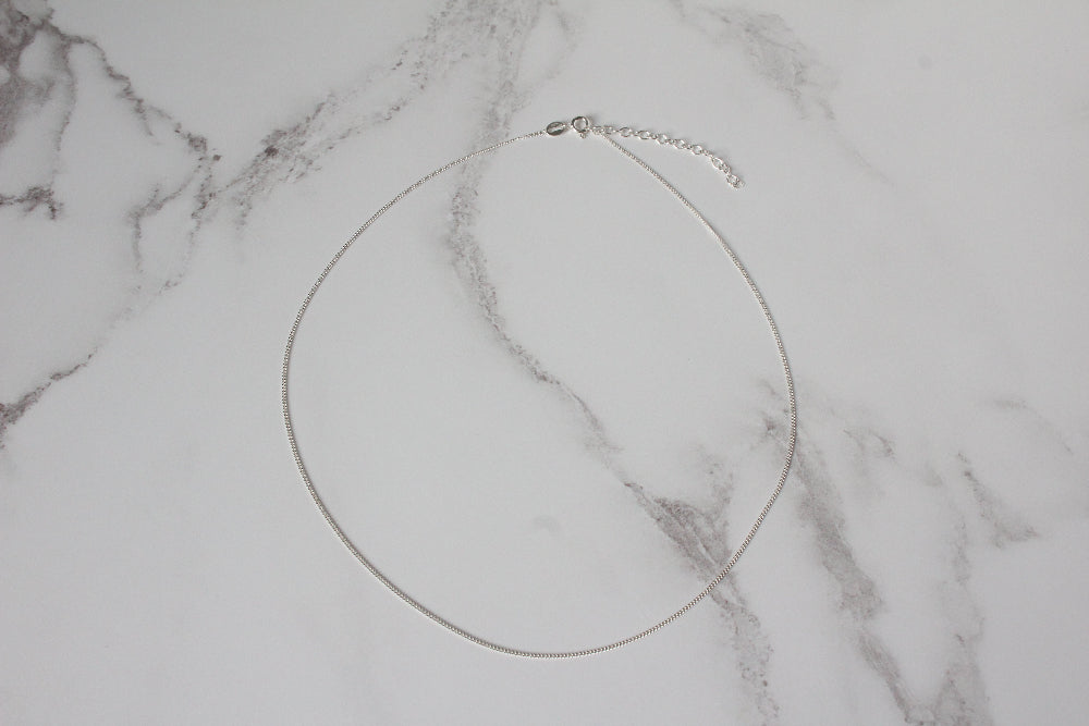 A delicate silver curb chain necklace with a schain extender, displayed on a white marble background.