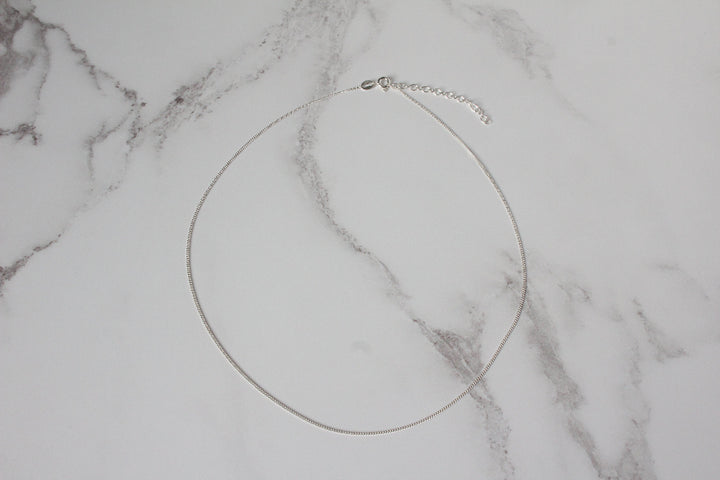 A delicate silver curb chain necklace with a schain extender, displayed on a white marble background.