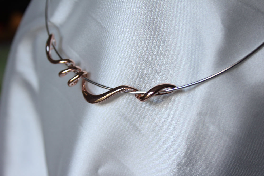 A close-up of a sculptural torque necklace featuring twisted rose gold piece and silver metal strands draped over a white satin fabric background.