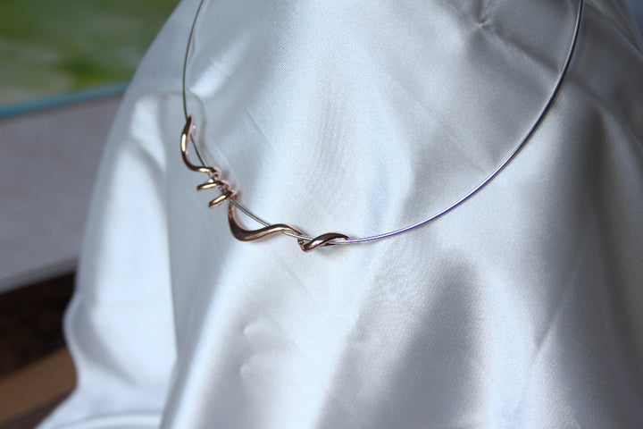 A silver torque necklace with rose gold twist draped on a white satin fabric.