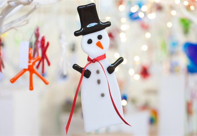 A white snowman Christmas ornament with a black top hat, black arms, a red ribbon necktie, and an orange carrot nose, hanging against a softly blurred festive background.