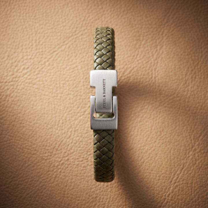 An olive green braided leather bracelet with a silver-tone metal clasp engraved with 'STEEL & MARNETT', displayed vertically on a textured tan leather background.