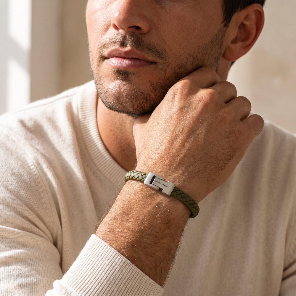 A close-up of a man wearing a green braided bracelet with a silver metal clasp, resting his hand on his chin.