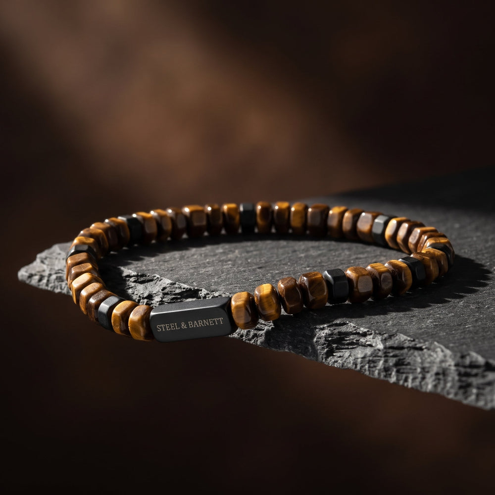 A beaded bracelet with alternating brown and black tiger's eye beads and a rectangular steel clasp engraved with 'STEEL & BARNETT', resting on a dark textured stone surface against a dark brown background.