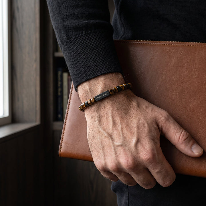 A close-up of a man's hand holding a brown leather folder, wearing a beaded bracelet with a central black metal tag engraved with 'steel & barnett'.