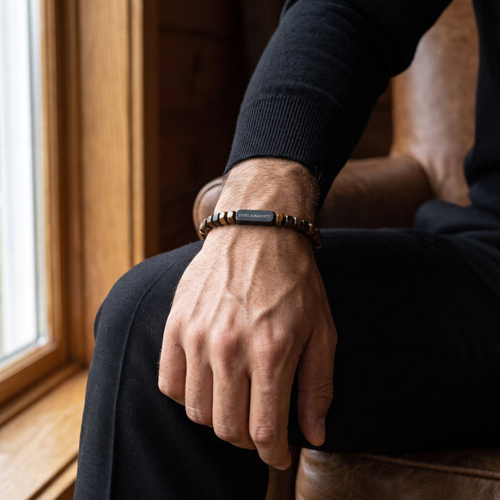 A close-up of a man's hand resting on his knee, wearing a beaded bracelet with a central black rectangular plate engraved with the brand name 'KEMPER'.