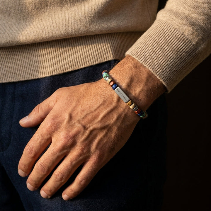 A close-up of a man's wrist wearing a personalized beaded bracelet with a silver metal nameplate engraved with 'JAMES'. The bracelet features multicolor beads and a silver-toned central plate.
