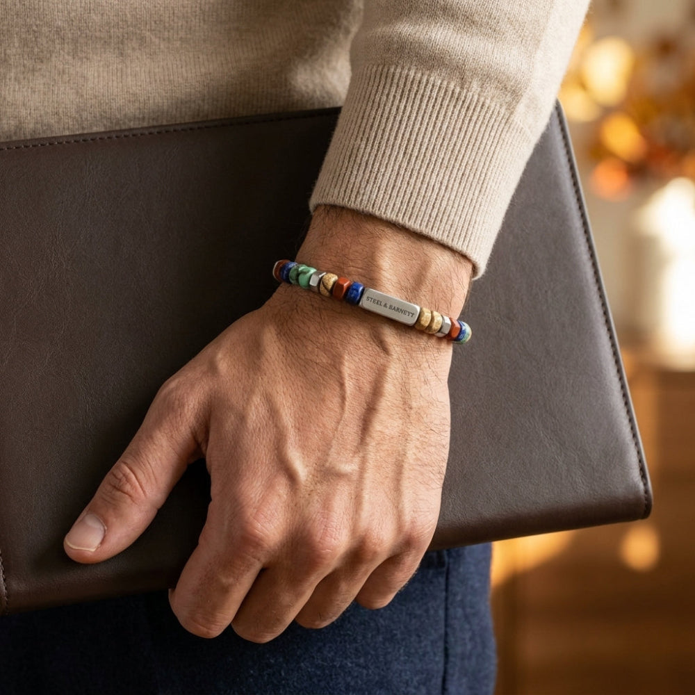 A close-up of a person's wrist wearing a colorful beaded bracelet with a silver metal tag engraved with 'Love Always'. The person is holding a dark brown leather folder, wearing a beige knit sweater, and blue jeans, with a warm, softly blurred background.