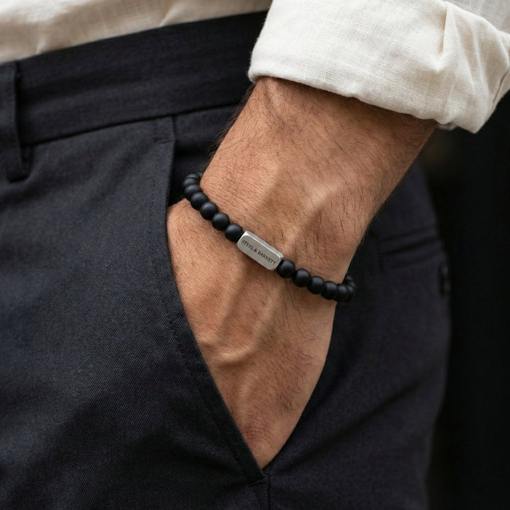 Black beaded bracelet with a metal tag on a wrist, worn with dark pants and a light shirt.