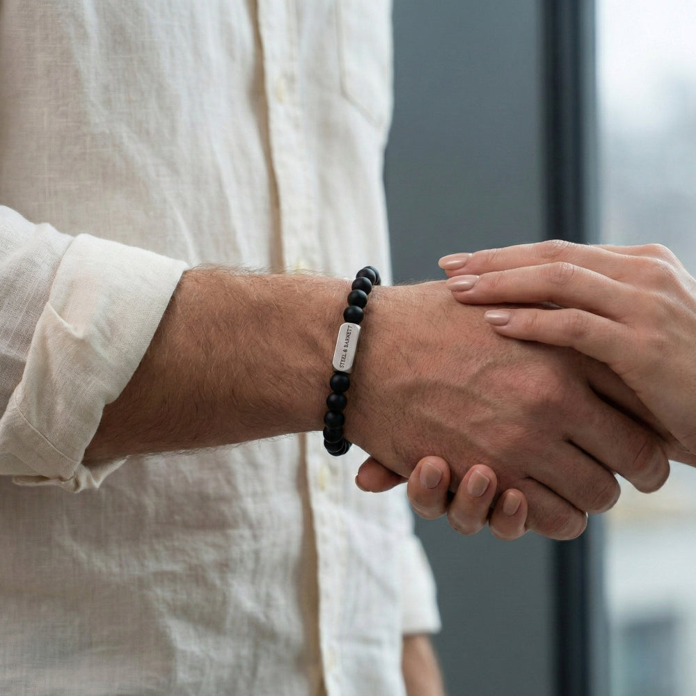 Two people shaking hands with one person wearing a black beaded bracelet with a silver tag.