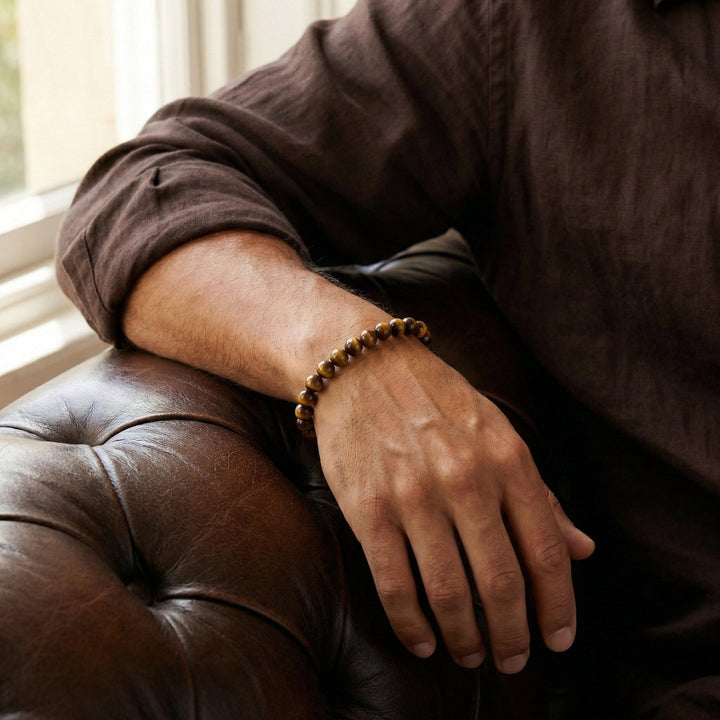 A close-up of a person's hand resting on a brown leather armchair, wearing a beaded bracelet made of brown and black tiger's eye stones.