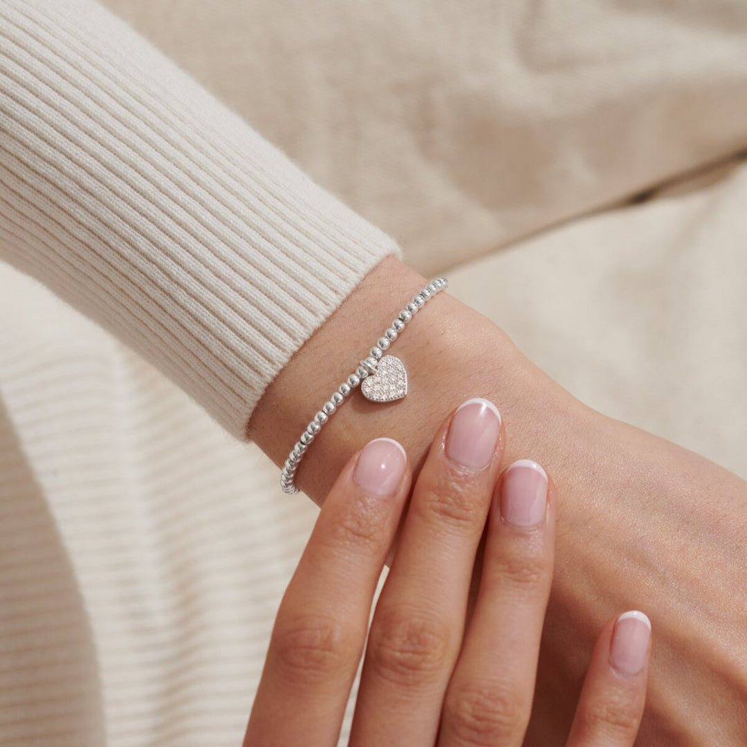 A close-up of a delicate silver chain bracelet with a small heart charm, worn on a woman's wrist. The bracelet is styled with a cream-colored ribbed knit sweater, and the background is a soft beige fabric.