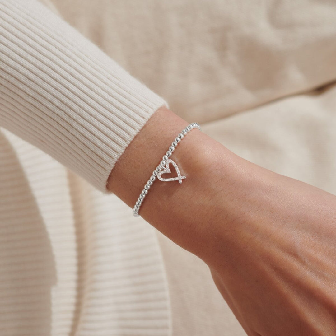A delicate silver chain bracelet with a small open heart charm, shown worn on a person's wrist over a cream ribbed sweater.